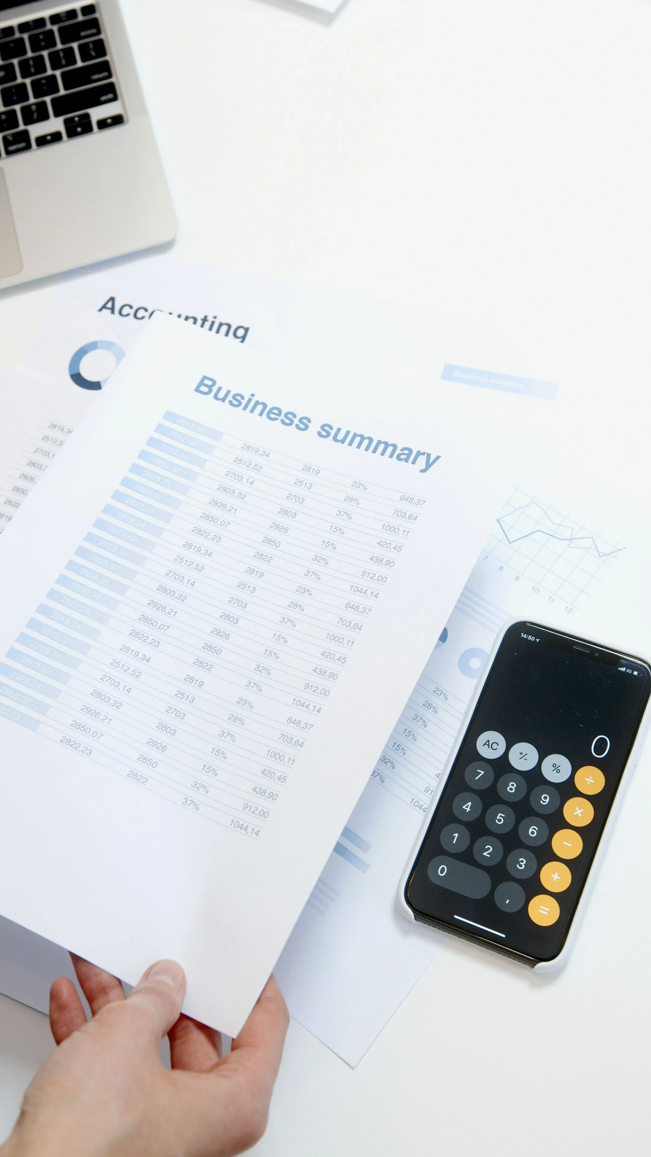 A hand holding printed accounting and business summary reports beside a calculator app open on a smartphone