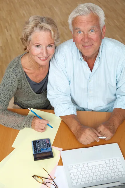 An elderly couple sit together at a table, working through paperwork with a calculator and laptop