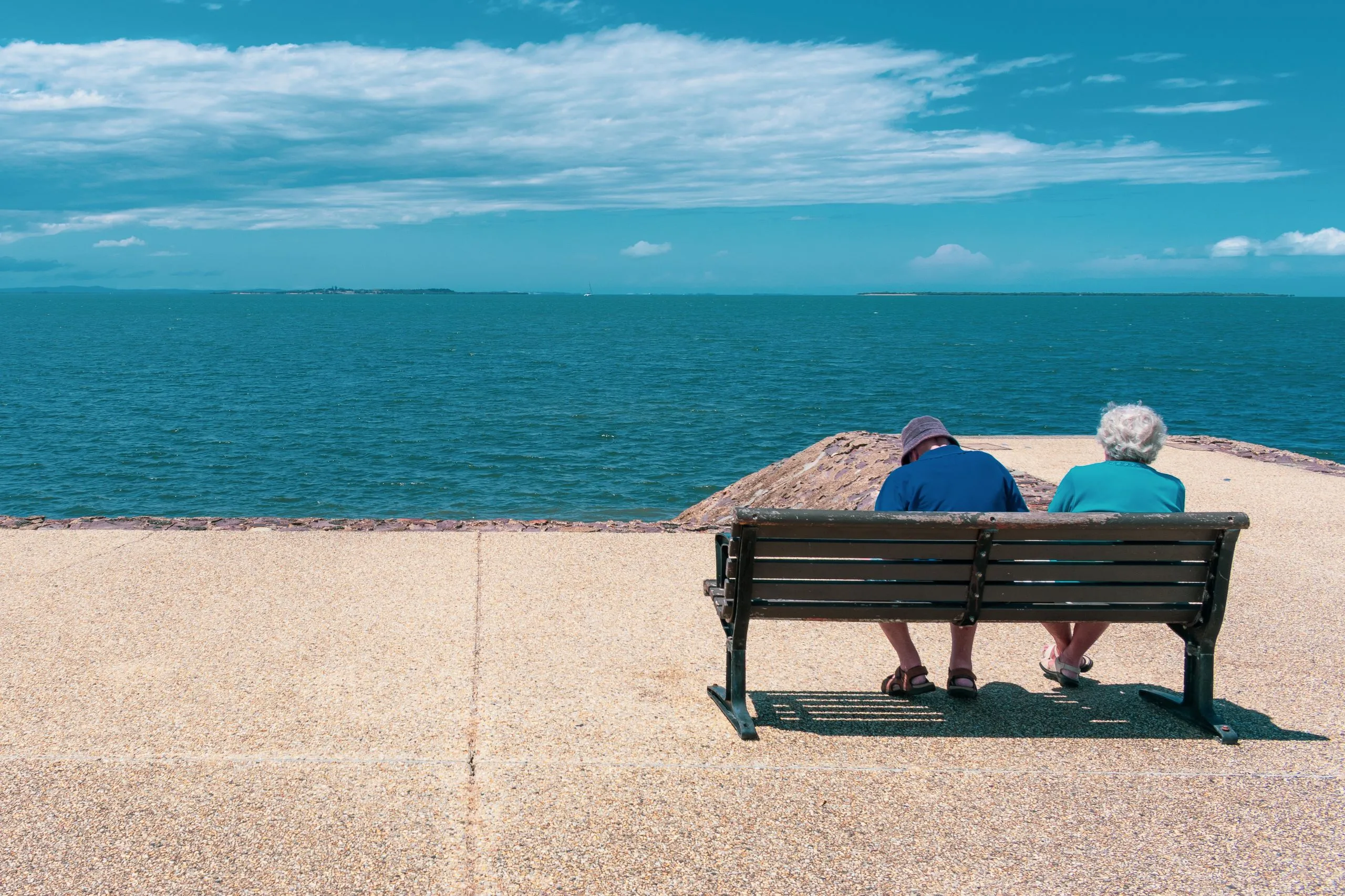 An older couple sitting on a bench by the water, viewed from behind, looking out at the sea.