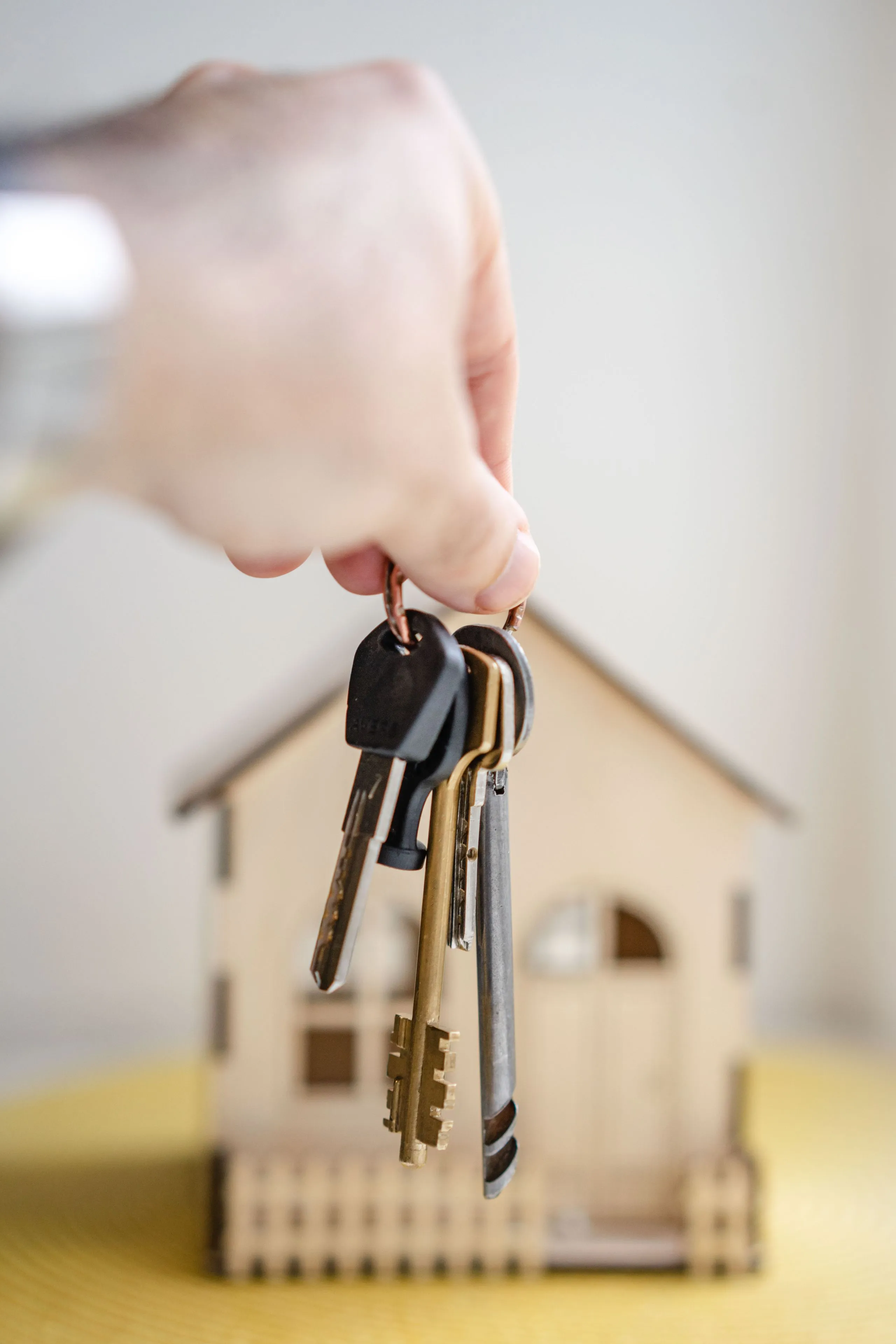 A hand holding a bunch of house keys in front of a small wooden model house on a yellow surface.