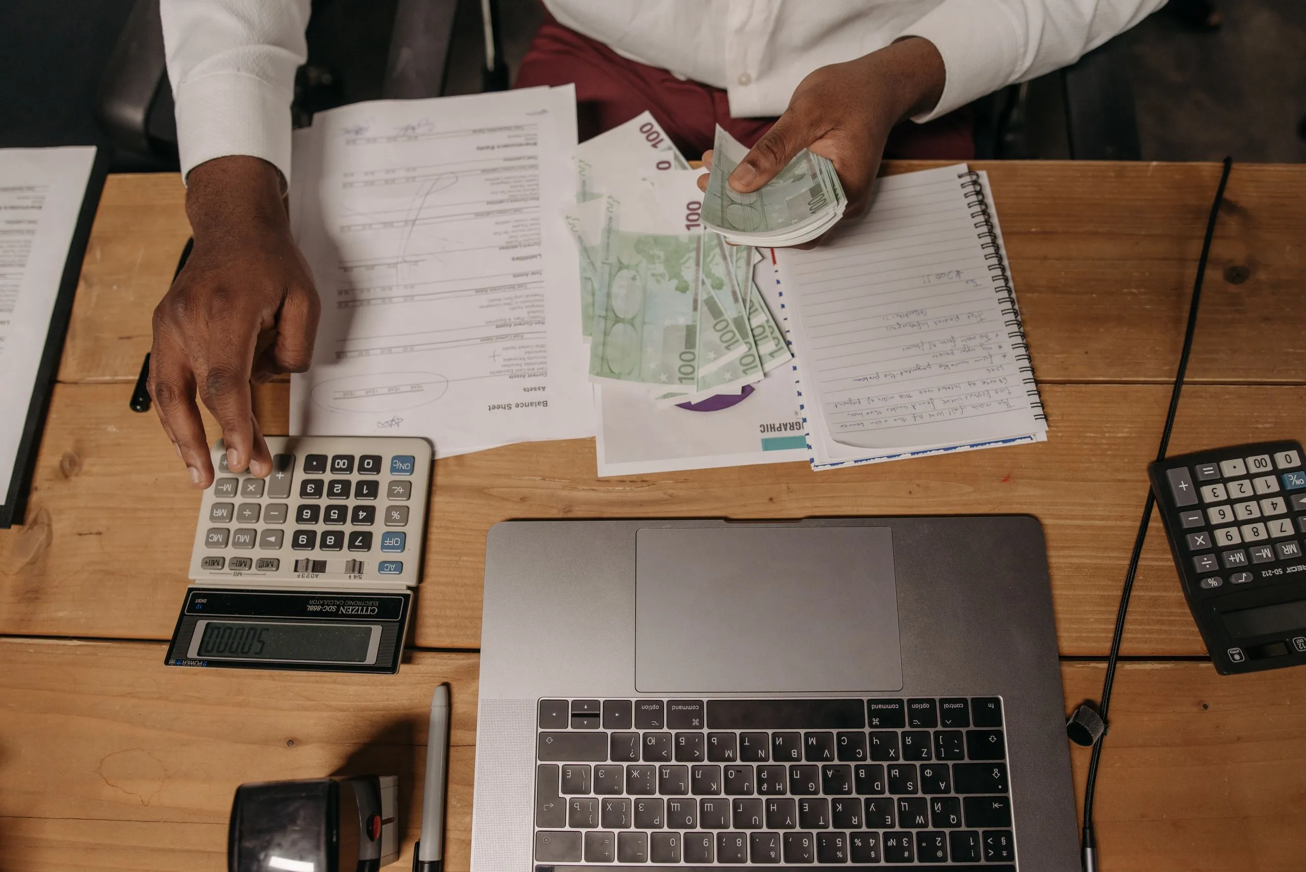 A person counting banknotes at a desk covered in financial paperwork, with a calculator and laptop nearby.