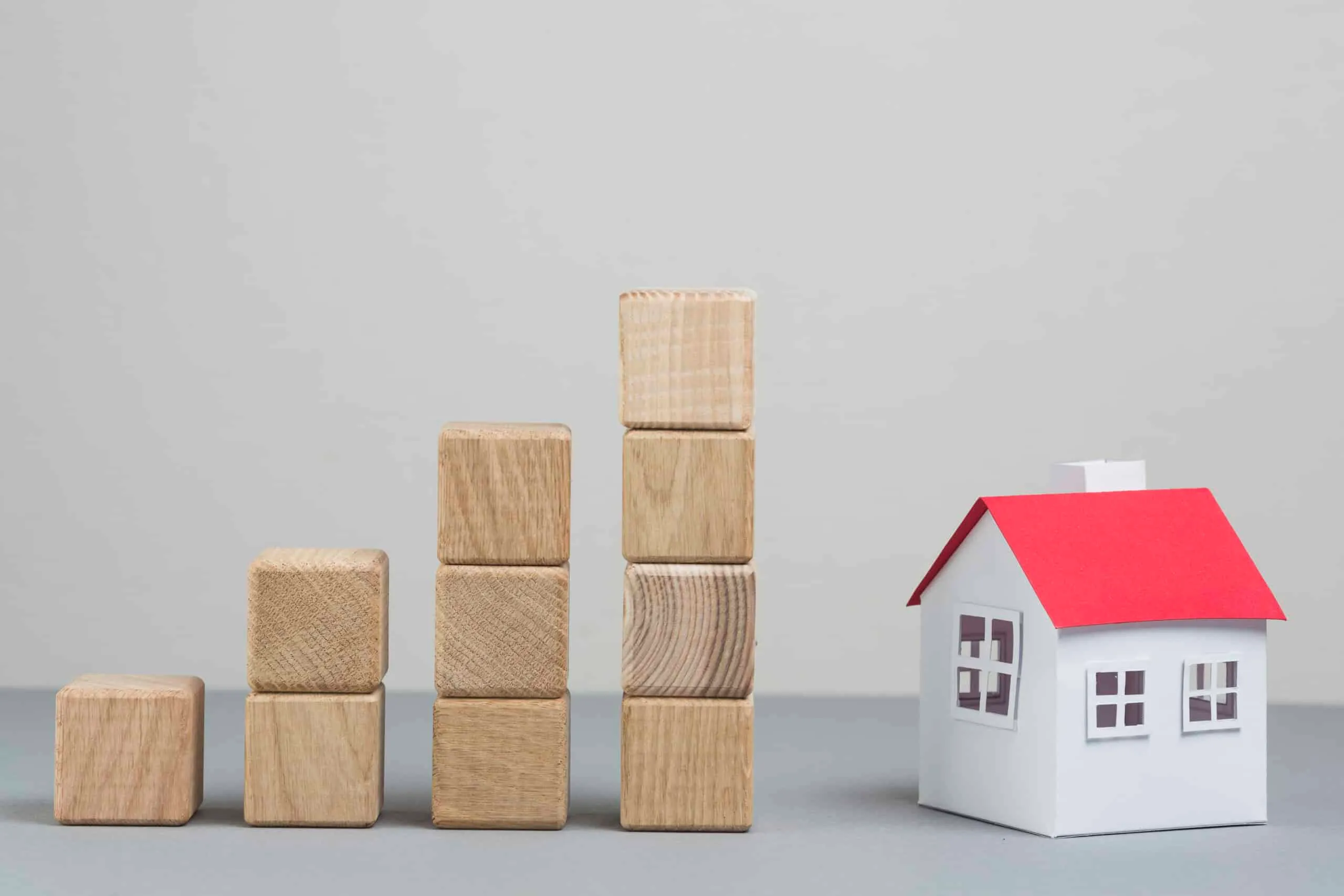 A small model house sits beside a stepped stack of wooden blocks rising in height, set against a plain grey background.